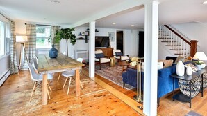 A spacious living area with a wooden dining table, white chairs, a blue sofa, and a staircase leading to another level.