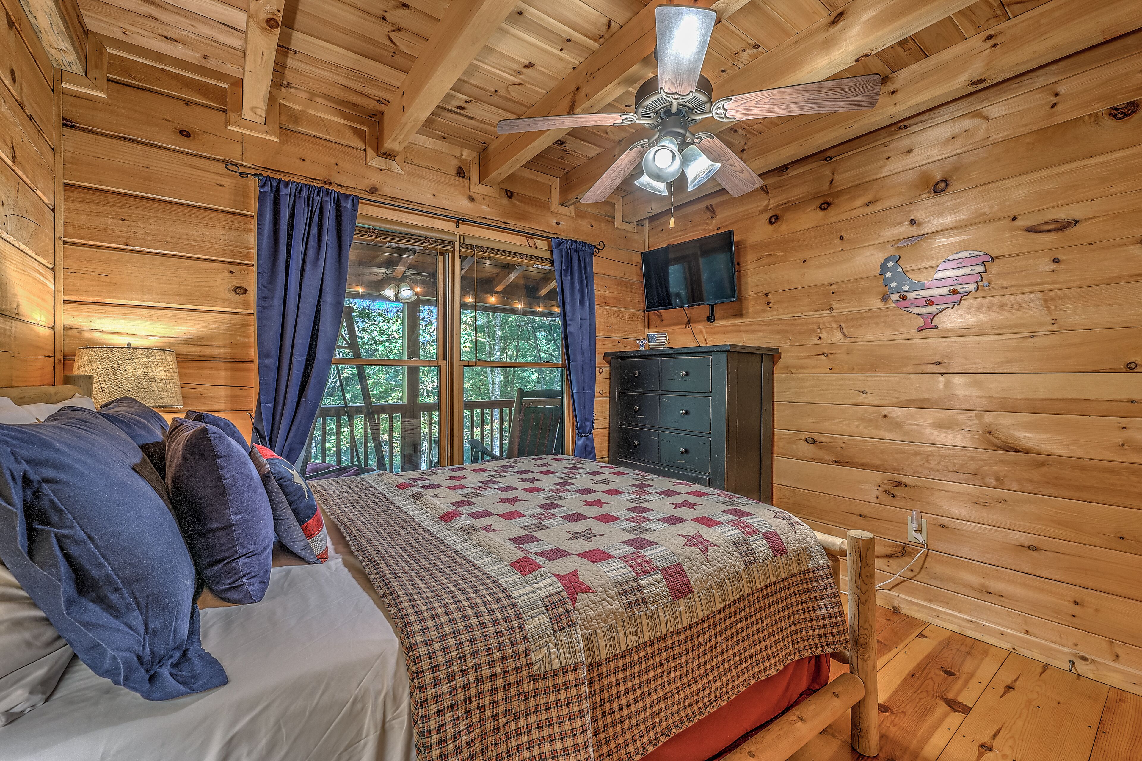 A wooden cabin bedroom with a bed, a ceiling fan, a dresser, and a television.