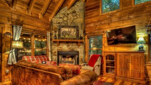 A cozy cabin interior with a stone fireplace, wooden walls, a large leather sofa, and a flat-screen TV. - Treetop 3br Cabin- Hot tub, Fireplace, Firepit, Grill, Fast Wi-Fi & Dog-Friendly (Blue Ridge)