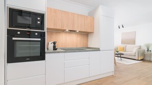 A modern kitchen with white cabinetry, a built-in oven, and a microwave. There is a wooden countertop and a small island with a sink. A living area with a sofa and a coffee table is visible in the background.