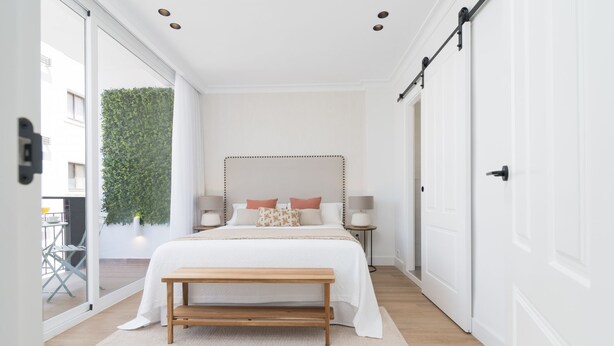 A bedroom with a large bed, bedside lamps, a bench, a sliding door, and a view of greenery outside.