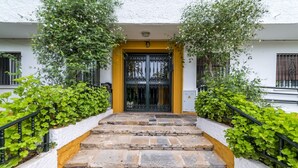 Un conjunto de escalones de piedra que conducen a una reja de metal negro, flanqueados por abundante vegetación verde y paredes blancas.