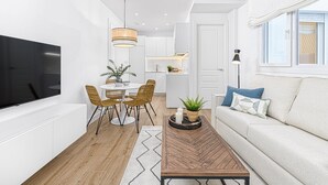 A modern living room with a flat-screen TV, a wooden coffee table, a sofa with cushions, and a dining area with wicker chairs and a white table.