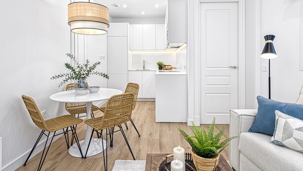 A modern living room with a white sofa, a dining table with wicker chairs, and a kitchen in the background.