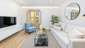 Una sala de estar moderna con una televisión de pantalla plana, un sillón azul, una mesa de centro de vidrio, un sofá gris y un espejo grande en la pared.