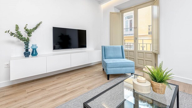 Una sala de estar moderna con un televisor de pantalla plana montado en un mueble blanco, un sillón azul, una mesa de centro de vidrio y un ventanal con vistas a un edificio.