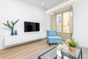 A modern living room with a flat-screen TV mounted on a white cabinet, a blue armchair, a glass coffee table, and a large window with a view of a building.