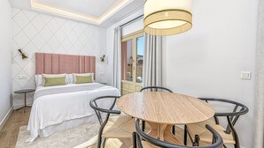 A hotel room with a bed, bedside tables, a round wooden table with chairs, a hanging light fixture, and a view through a glass door.