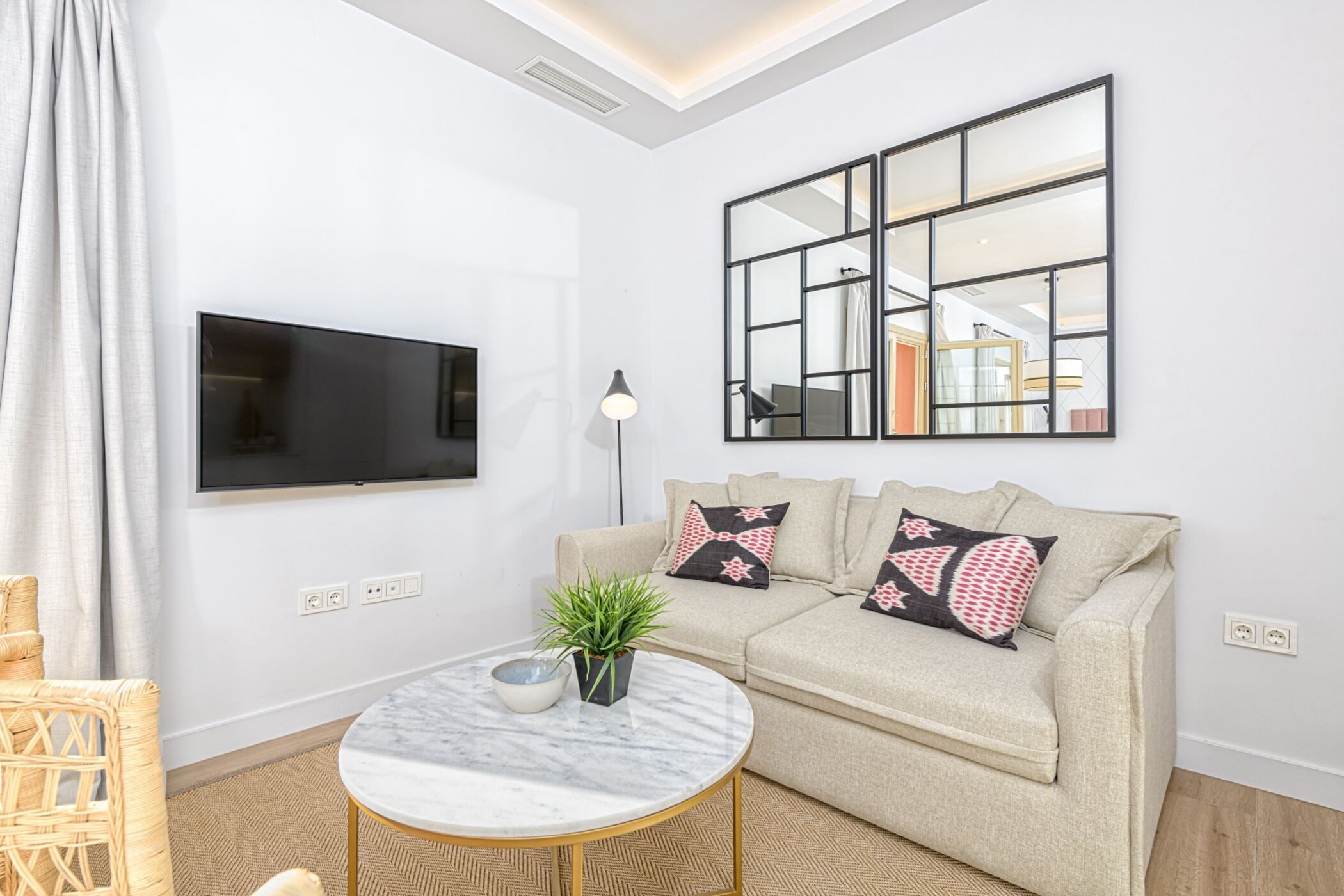 A modern living room with a flat-screen TV, a round marble coffee table, a beige sofa with patterned cushions, and a large mirror on the wall.