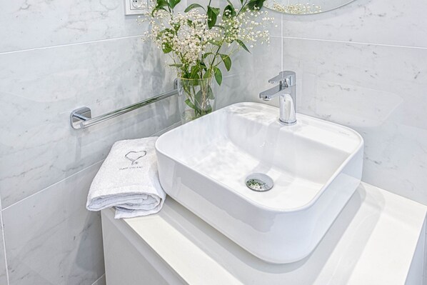 Un lavamanos blanco con grifo cromado, un florero de vidrio con flores blancas y una toalla blanca enrollada con un diseño de corazón.
