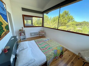 A bedroom with a large window, a bed with a colorful quilt, a small bedside table, and a chair.