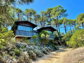 A two-story wooden cabin with a balcony, surrounded by dense pine trees and a dirt path leading to the entrance. - Architect villa in nature near ocean & bay A0389 (Lège-Cap-Ferret)