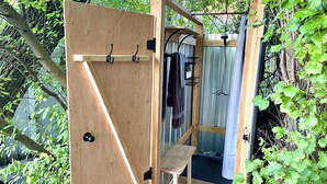 A wooden outdoor shower area with a bench, hooks for hanging towels, and a metal shower head. - Glamping | Outdoor Shower | Bathroom (Providence)