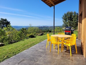An outdoor patio with a round table and yellow chairs, a barbecue grill, and a view of the countryside. - Villa Celeste Ardeche (UCEL)