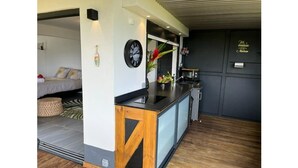 A modern kitchen with a black countertop, wooden cabinets, and a built-in oven. There is a dining area with a table and chairs, and a bed with a patterned bedspread in the background. - Studio O'KALM - Le Gosier (LE GOSIER)