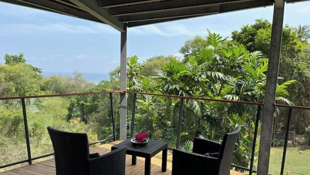 A covered outdoor area with two wicker chairs and a small table, surrounded by lush greenery and a view of the ocean. - Studio O'KALM - Le Gosier (LE GOSIER)