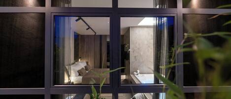 A modern interior with a large glass window, a bed, a bedside table, and a bathroom visible through the window.