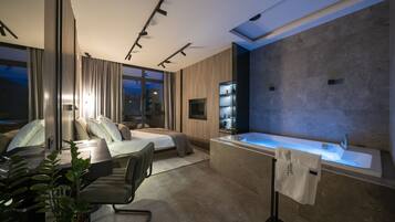 A modern hotel room with a large bed, a sitting area with a chair, a desk, a potted plant, a large window, and a bathroom with a bathtub and sink.