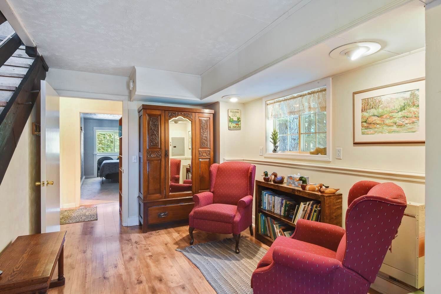 A cozy living room with a wooden floor, a red armchair, a matching ottoman, a wooden bookshelf, and a large mirror.