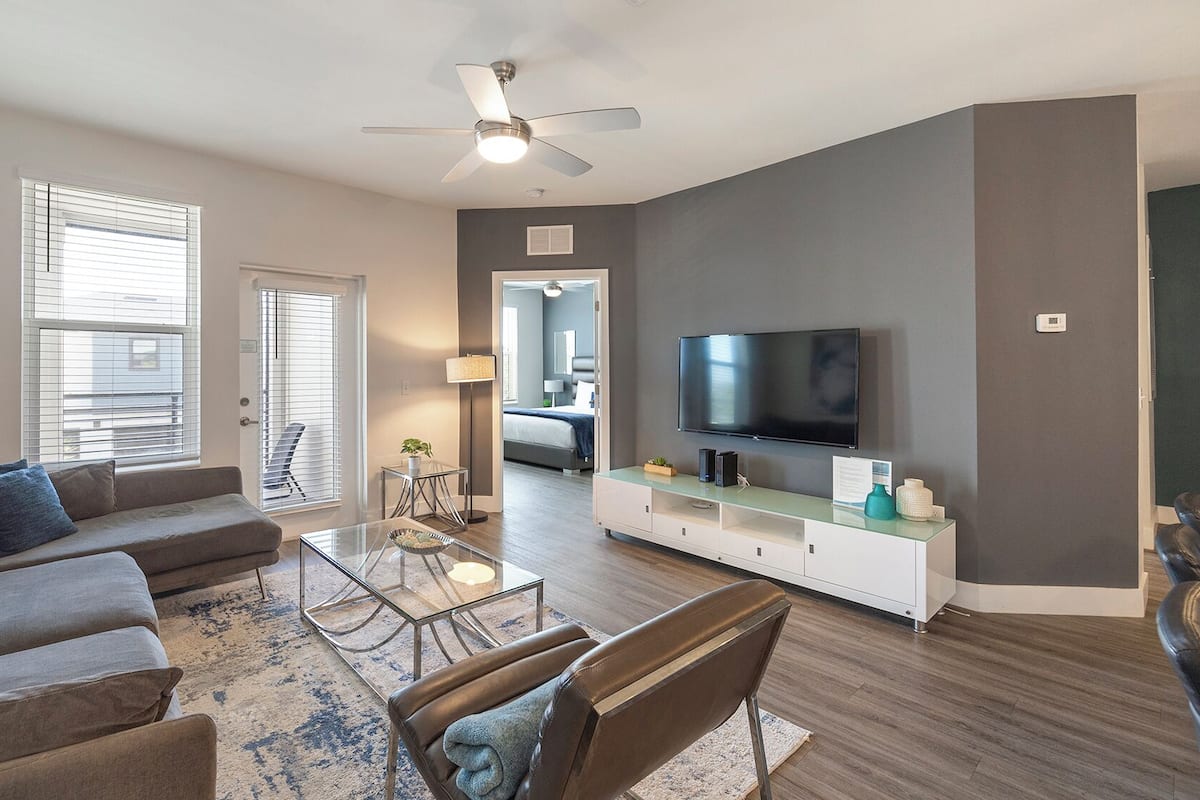 A modern living room with a sofa, coffee table, and TV. There is a ceiling fan, a window with blinds, and a door leading to another room.