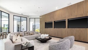 A modern living room with a sectional sofa, a coffee table, and a large gray pouf. There are three flat-screen TVs mounted on a wooden wall. - Landing Tampa Westshore (Tampa)