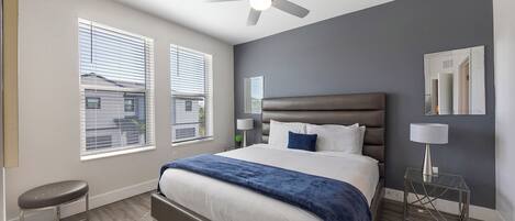 Ein Schlafzimmer mit einem großen Bett, einem Deckenventilator, zwei Fenstern mit Jalousien, einem Nachttisch und einem Beistelltisch.