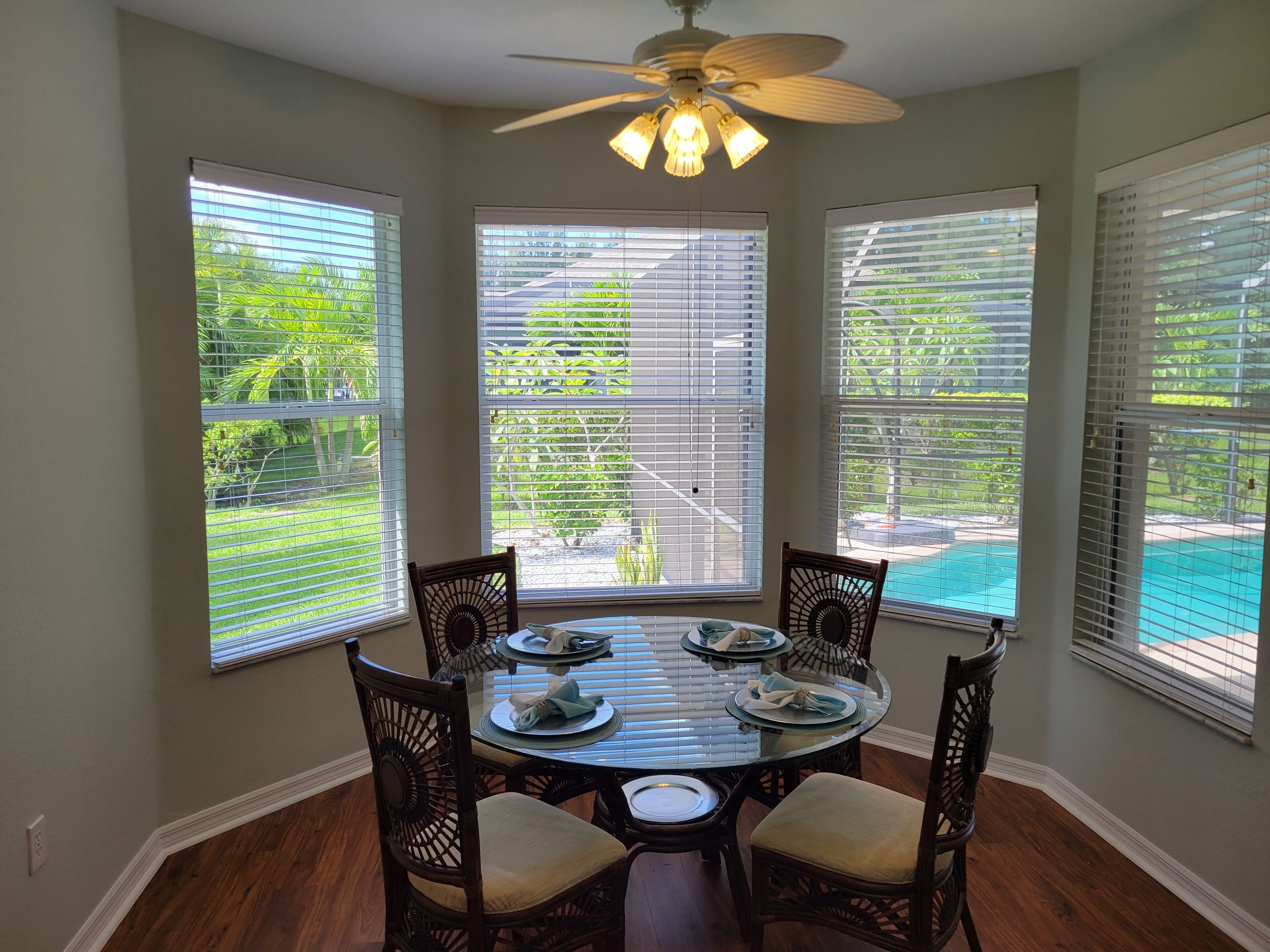 A dining area with a round table, four chairs, and large windows with blinds.