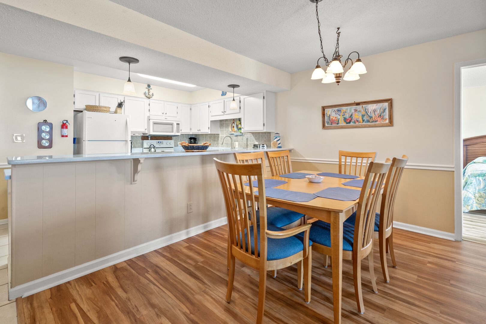 A modern kitchen with white cabinets, a central island, and a dining area with wooden chairs and blue cushions.