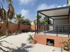 A covered outdoor seating area with a gravel floor, surrounded by palm trees and potted plants.