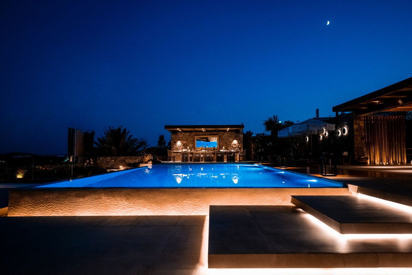 Vista noturna de uma piscina com uma estrutura de pedra ao fundo, cercada por degraus e caminhos bem iluminados.