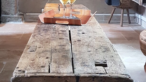 Een houten tafel met een rustieke uitstraling, waarop twee glazen witte wijn, een kaars en een kussen in de vorm van een ster staan. Op een houten bankje.