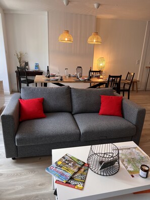 A modern living room with a grey sofa, red cushions, a coffee table with magazines and a candle, and a dining area in the background.