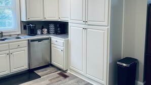 A kitchen with white cabinets, a coffee maker, a coffee pot, and a trash can. - Historic Pursuit of Happyness (Gastonia)