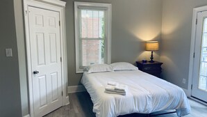 A bedroom with a bed, a nightstand, a lamp, and a window with blinds. - Historic Pursuit of Happyness (Gastonia)