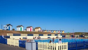 Ein Schwimmbeckenbereich mit Holzzaun, Rettungsschwimmerstand und bunten Strandhäusern im Hintergrund.