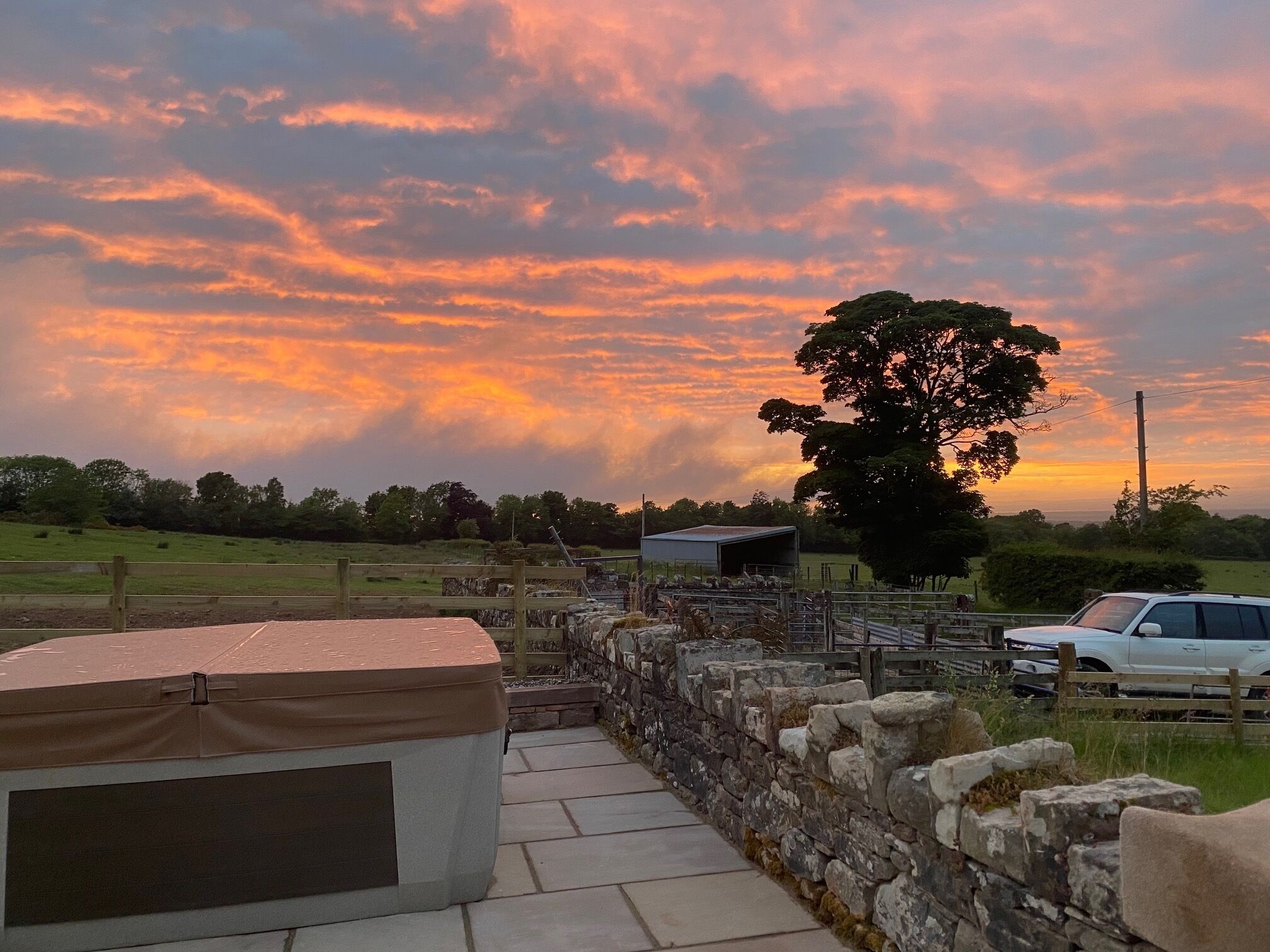 A stone wall with a covered bench, a fenced area with animals, and a tree in the background.