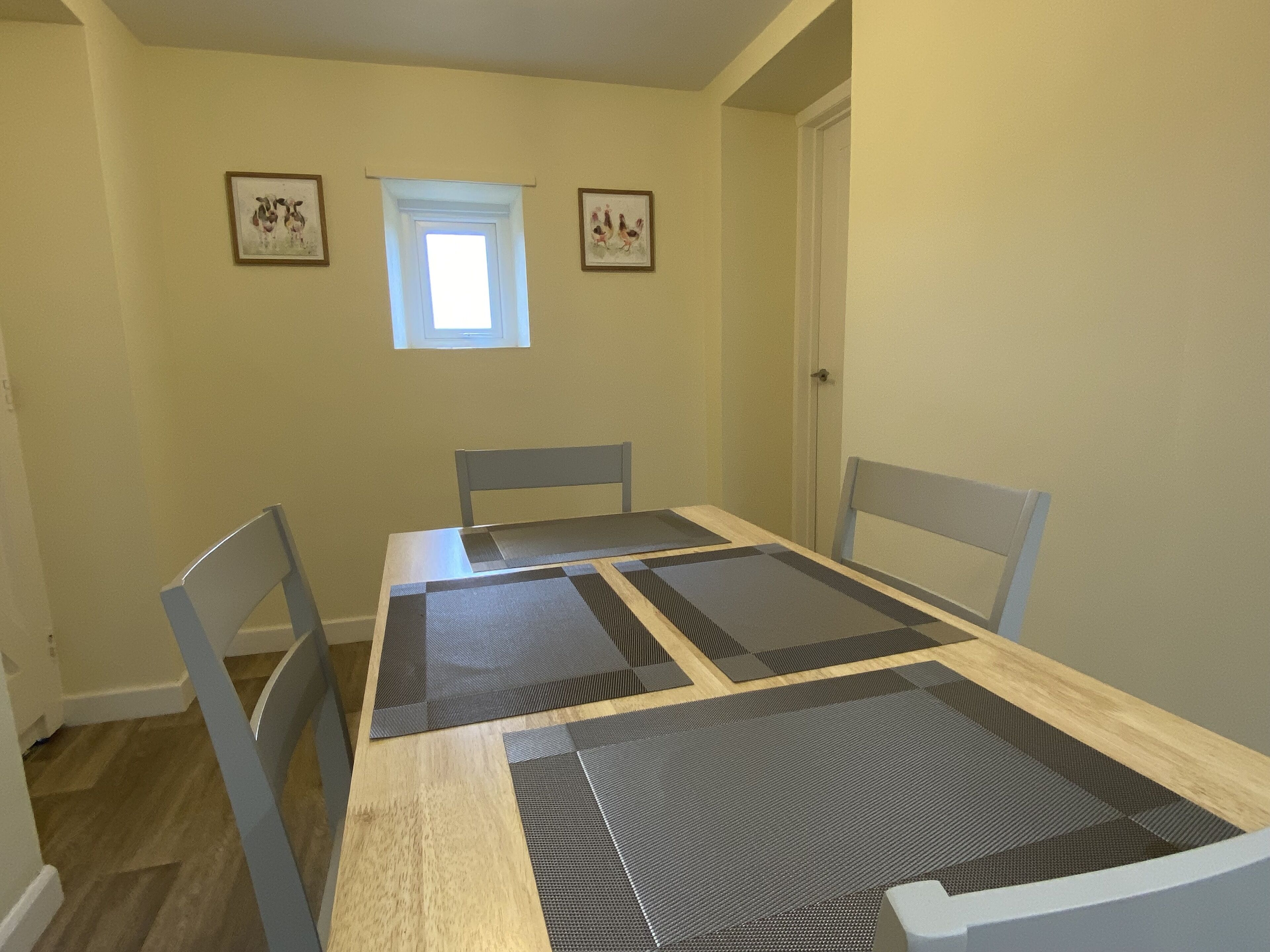 A dining room with a wooden table, four chairs, two framed artworks, and a small window.