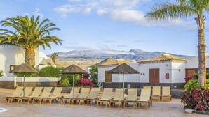 Ein Poolbereich mit Liegestühlen und Sonnenschirmen, umgeben von Palmen und einer Bergkulisse.