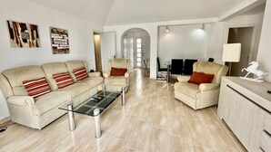 Ein geräumiges Wohnzimmer mit hoher Decke, einem Glastisch, einer beigefarbenen Sofagarnitur und einem Essbereich mit Stühlen und Tisch.