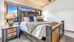 A bedroom with a wooden ceiling, a bed with a rustic headboard, a nightstand with a lamp, and a view through large windows. - Brand New Construction! Ski-In/Ski-Out Townhome with Private Hot Tub! (Whitefish)