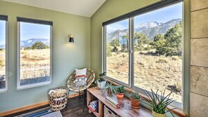 Ein Zimmer mit großen Fenstern und Blick auf die Berge, ein Korbstuhl, ein kleiner Tisch mit Pflanzen und einer Wandleuchte.