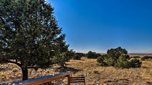 Eine Holzterrasse mit Bank und Tisch, mit Blick auf eine trockene Landschaft mit vereinzelten Bäumen und blauem Himmel.