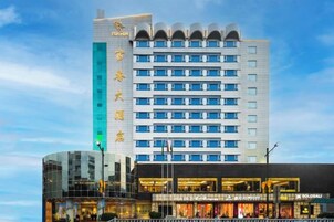 Un hotel de varios pisos con diseño moderno, que destaca un letrero vertical con texto, y una zona comercial a nivel de calle con tiendas.