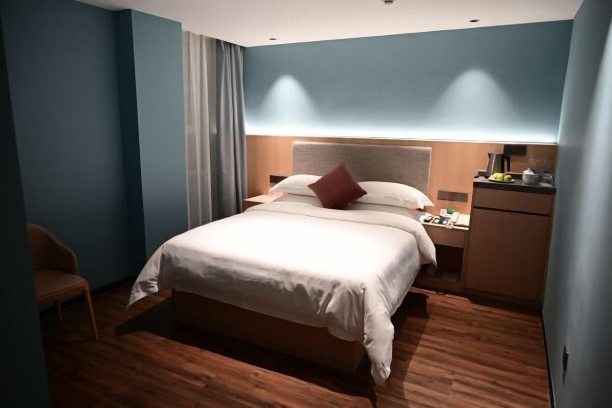 A modern hotel room with a large bed, a nightstand, a desk with a lamp, and a wardrobe.