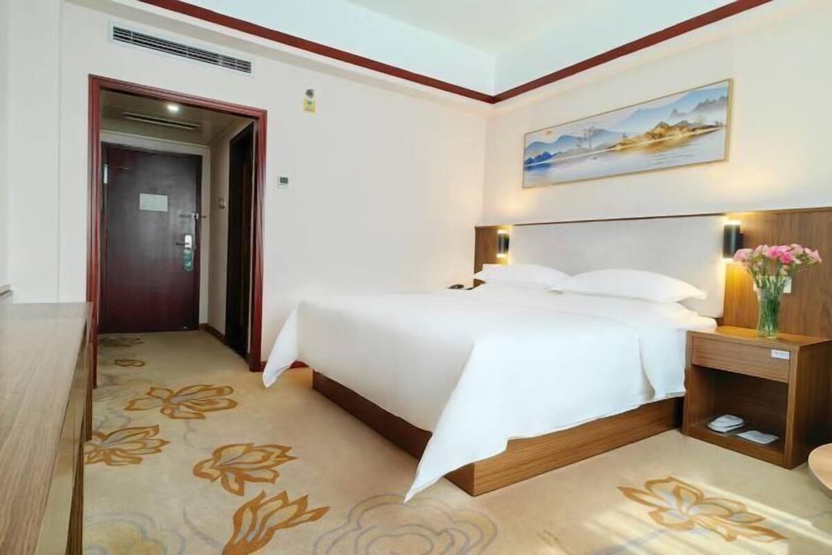 A hotel room with a bed, wooden headboard, bedside table with a vase, and a painting on the wall.