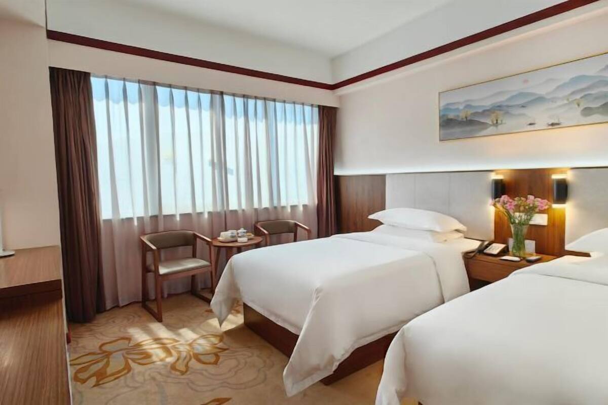 A hotel room with two beds, a desk with a chair, a painting on the wall, and a window with curtains.