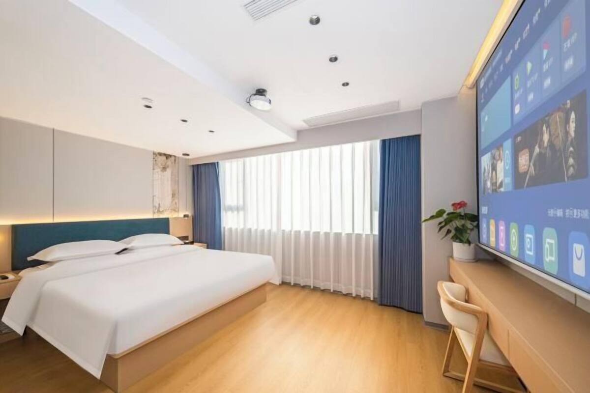 A modern hotel room with a large bed, a desk, and a large screen displaying room information.