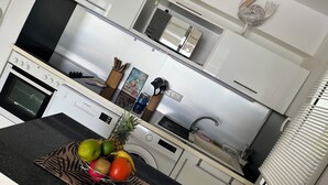 Moderni keittiö, jossa on sisäänrakennettu uuni, mikroaaltouuni ja ruostumattomasta teräksestä valmistetut kodinkoneet. Työtasolla on hedelmäkulho ja taustalla näkyy pesukone.