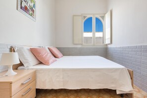 Une chambre à coucher comprenant un lit, une table de chevet, une lampe, une fenêtre avec un volet ouvert et une œuvre d’art accrochée au mur.