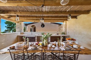An outdoor dining area with a wooden table set for a meal, surrounded by chairs, and a rustic ceiling with hanging light fixtures.